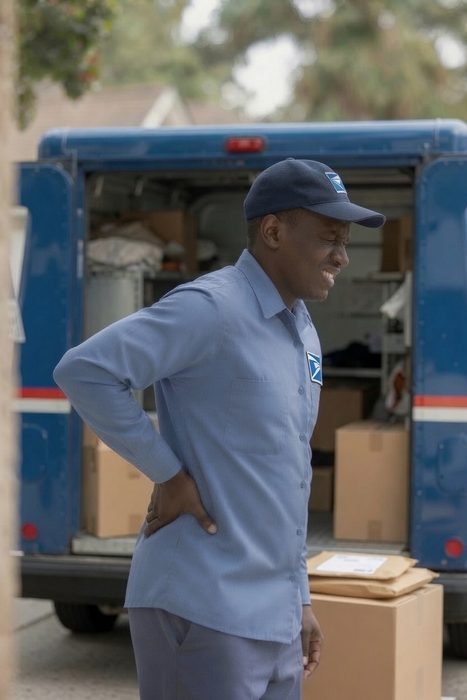 A USPS worker outside his delivery truck holding his lower back in pain.